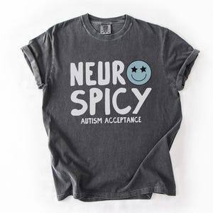May include: A dark gray t-shirt with the text "Neuro Spicy" and the words "Autism Acceptance" printed in white. A blue smiley face with two stars for eyes is above the text.