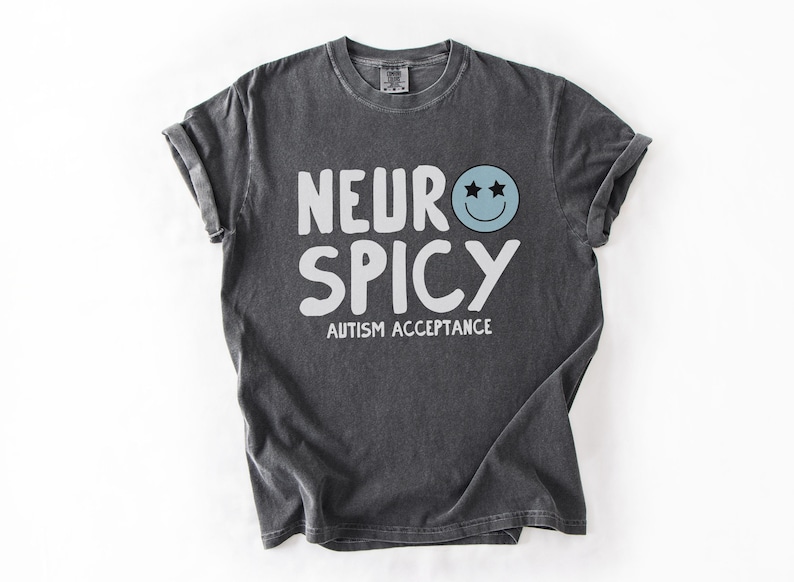 May include: A dark grey t-shirt with the text "Neuro Spicy" and the words "Autism Acceptance" printed in white. A blue smiley face with two stars for eyes is above the text.