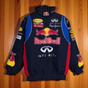 Op de afbeelding: Marineblauwe jas met rode, blauwe en gele accenten. De jas heeft het Red Bull-logo en andere sponsorlogo's. Het heeft een hoge kraag en een ritssluiting. De jas hangt aan een metalen hanger tegen een houten achtergrond.