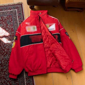 Puede incluir: Chaqueta de carreras roja con una franja negra en el pecho y detalles blancos. La chaqueta tiene un forro acolchado y un cuello alto. La chaqueta está abierta, revelando el interior. Sobre un suelo de madera.