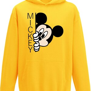 Puede incluir: Sudadera amarilla con capucha con una imagen de dibujos animados en blanco y negro de Mickey Mouse asom&aacute;ndose por una esquina. El texto "MICKEY" est&aacute; impreso verticalmente en el lado izquierdo de la imagen.