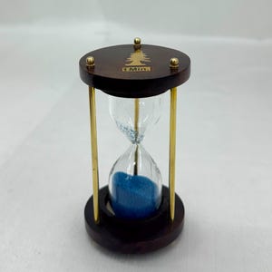 May include: An hourglass with a dark brown wooden frame and brass-colored metal supports. The glass bulb contains blue sand, and the top is marked with "1 Min." and a tree design. The hourglass is a decorative item.