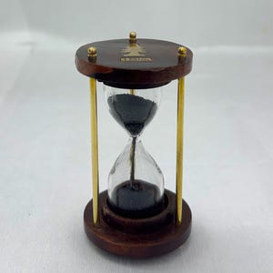 May include: An hourglass with a dark wood frame and base, featuring a clear glass chamber filled with black sand. The top and bottom are connected by gold-colored metal rods. The top has a small tree design and the word "TIME".