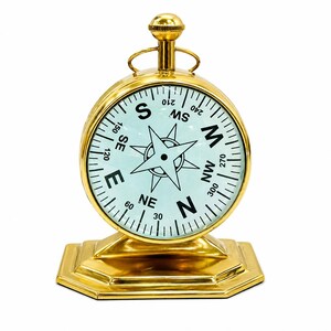 May include: A gold-colored compass with a white face and black lettering. The compass rose is in the center, and the cardinal directions are labeled. The compass sits on a gold-colored, octagonal base.