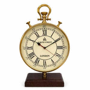 Op de afbeelding: Een klok in vintage-stijl met een crèmekleurige wijzerplaat, zwarte Romeinse cijfers en gouden wijzers. De klok leest "49 BOND STREET LONDON" en staat op een donkerbruine houten basis. Hij heeft een gouden handvat en twee kleine knoppen.
