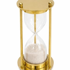 May include: An hourglass featuring a polished gold-tone metal frame with circular top and bottom plates. The frame is supported by three vertical rods. The clear glass bulb contains fine, light-coloured sand, with the majority settled at the base.