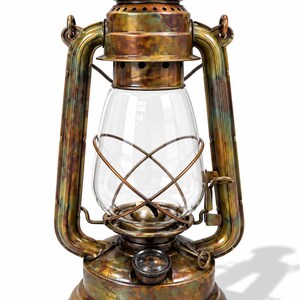 Może przedstawiać: Lampa naftowa z patynowanym, wielokolorowym wykończeniem w odcieniach brązu, miedzi i zieleni. Lampa posiada przezroczysty szklany klosz chroniony metalową klatką z drutu, okrągłą podstawę oraz górny uchwyt do przenoszenia lub zawieszania.