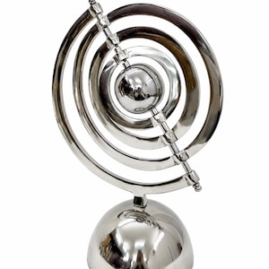 May include: A polished silver metal sculpture with concentric circular rings and a central sphere. The sculpture is mounted on a rounded base and features a diagonal rod with connected spheres. The design is abstract and modern.