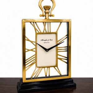 May include: A gold-colored rectangular clock with Roman numerals and black hands, set on a black base. The clock face is white with the text "McLaughlin & Scott ESTD 1972". The clock has a decorative handle at the top.