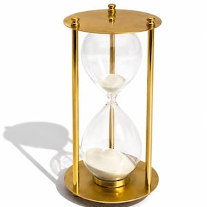 May include: An hourglass featuring a gold-toned metal frame with circular top and bottom plates connected by three vertical rods. The central glass bulb contains white sand, with a small portion currently resting in the top chamber.