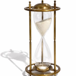 May include: An hourglass featuring a polished brass frame with a circular base and top. The clear glass bulb contains fine white sand, with the majority of the sand currently held in the upper chamber, supported by four vertical brass pillars.