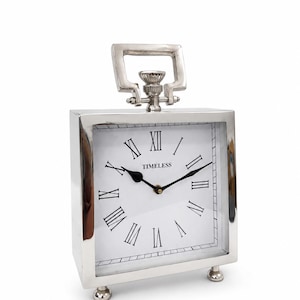 Puede incluir: Un reloj de mesa cuadrado de metal plateado con esfera blanca, números romanos negros y manecillas negras. La palabra TIMELESS está impresa en negro en la esfera. El reloj cuenta con un asa superior y dos pequeños pies esféricos en la base.