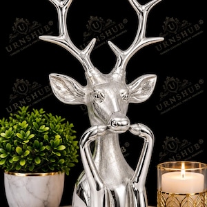 May include: A silver-colored deer figurine featuring a detailed head with large antlers and front legs positioned near the snout. The sculpture has a polished, reflective metallic finish and is presented in a vertical, upright orientation.
