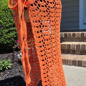 May include: A bright orange crochet wrap skirt featuring an intricate openwork shell stitch pattern. The garment includes a long, integrated tie closure at the waist, allowing for an adjustable fit. The design creates a semi-sheer, textured appearance.