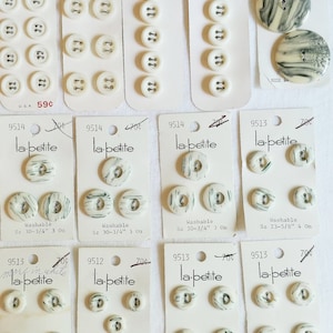 May include: A collection of white plastic buttons with green and black designs. The buttons are packaged on cards with the brand name "la petite" and size information. The cards also include the text "Washable" and "more in white".