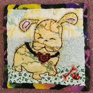 May include: A quilted fabric square with a hand-stitched image of a brown and yellow dog wearing a black collar with a red heart. The dog is standing on a green and white fabric with a bone and a butterfly.