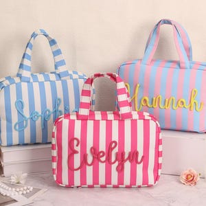 Op de afbeelding: Drie gestreepte draagtassen in blauw en wit, roze en wit en roze en blauw. Elke tas heeft een andere naam geborduurd: "Sophie", "Evelyn" en "Hannah". De tassen hebben handvatten en staan op een wit oppervlak.