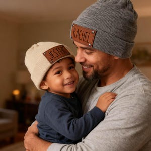 May include: A father and child are embracing, both wearing personalized beanies. The child's beanie is cream-colored with the name "JESTER" on a brown leather patch. The father's beanie is gray with the name "RACHEL" on a similar patch.