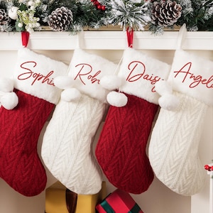 Puede incluir: Cuatro calcetines navideños personalizados. Dos son rojos con la parte superior blanca y dos son blancos con la parte superior blanca. Cada calcetín tiene un nombre bordado en escritura roja. Una guirnalda festiva con piñas y bayas está arriba.
