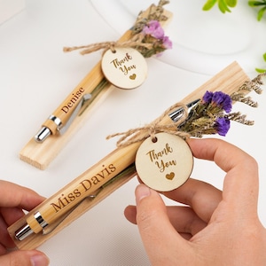 May include: Two personalised pens with wooden accents, each in a wooden holder. The pens are engraved with names and have a small wooden tag saying "Thank You" with a heart. Dried purple flowers and twine complete the design, ideal for gifting.