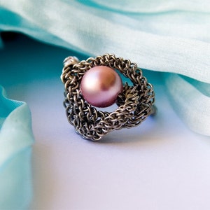 Könnte beinhalten: Ein silberner Ring mit einer rosa Perle in der Mitte. Der Ring ist aus einem gewebten Metalldesign gefertigt.