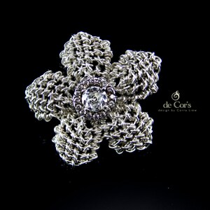 Peut inclure: Une broche en forme de fleur argentée avec un strass transparent au centre. La broche est faite de métal tissé complexe.