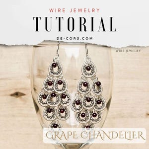 May include: Silver wire earrings with a grape chandelier design. The earrings feature multiple layers of teardrop-shaped wirework with small, dark red beads. The earrings are displayed against a wooden background.