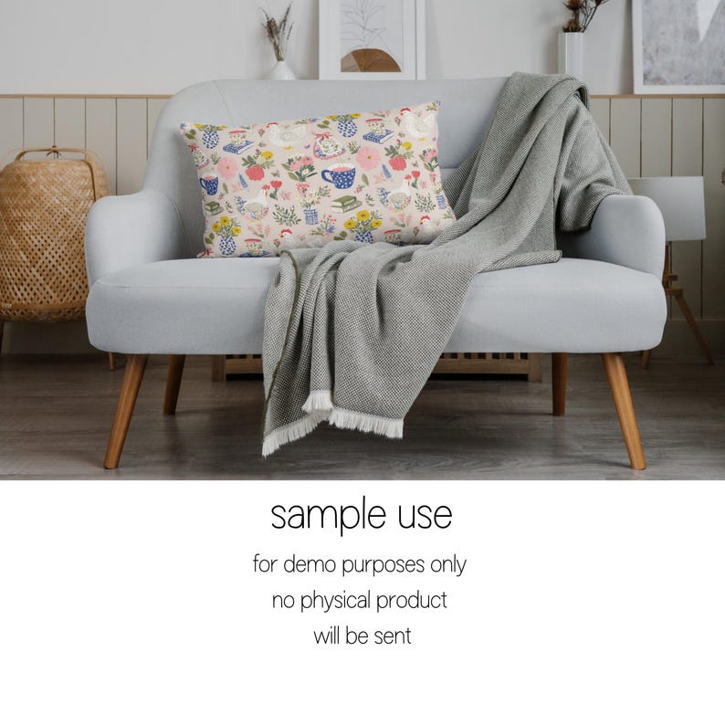 May include: A patterned cushion with a tea party design sits on a light grey sofa. The cushion displays teacups, flowers, and books in pink, blue, and yellow. A grey blanket with a fringed edge is draped over the sofa, which has wooden legs.