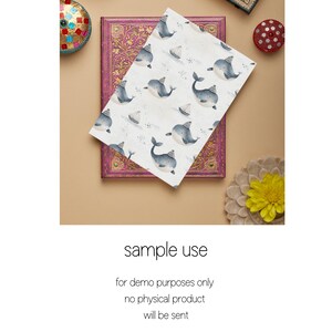 May include: A white card with a repeating pattern of blue whales wearing party hats and small sailboats. The card is lying on top of a closed book with a pink and gold cover.