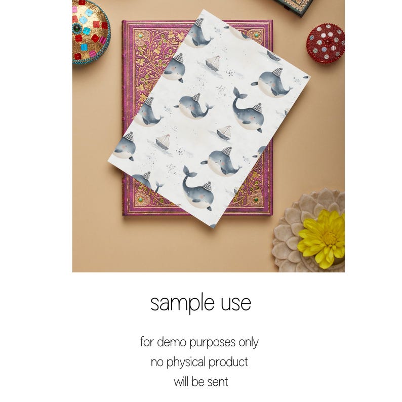 May include: A white card with a repeating pattern of blue whales wearing party hats and small sailboats. The card is lying on top of a closed book with a pink and gold cover.