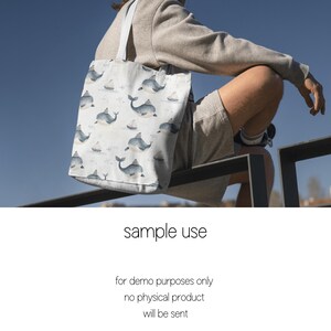 May include: A white tote bag with a blue whale pattern. The bag is being held by a person wearing a grey sweater and shorts.