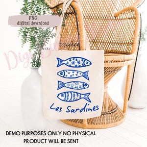 May include: A beige tote bag featuring four blue fish illustrations with decorative patterns. The words "Les Sardines" are written in blue script below the fish. The bag is set against a wicker chair and white vase.