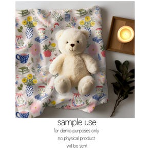 May include: A cream teddy bear rests on a floral blanket featuring vases, books, and teacups. The blanket has white, pink, yellow, and blue details. A lit candle in a wooden tray and greenery are also visible.