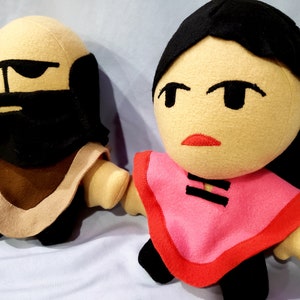 May include: Two plush toys, one with a brown beard and a tan robe, the other with black hair and a pink robe with black trim.