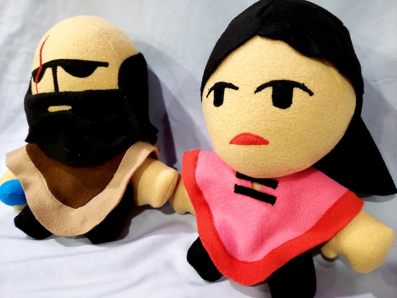 May include: Two plush toys, one with a brown beard and a tan robe, the other with black hair and a pink robe with black trim.