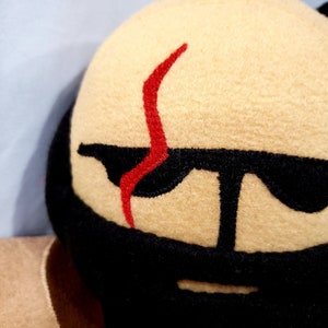 May include: A plush toy with a tan face, black beard, and red scar. The toy has black eyes and a black mouth.