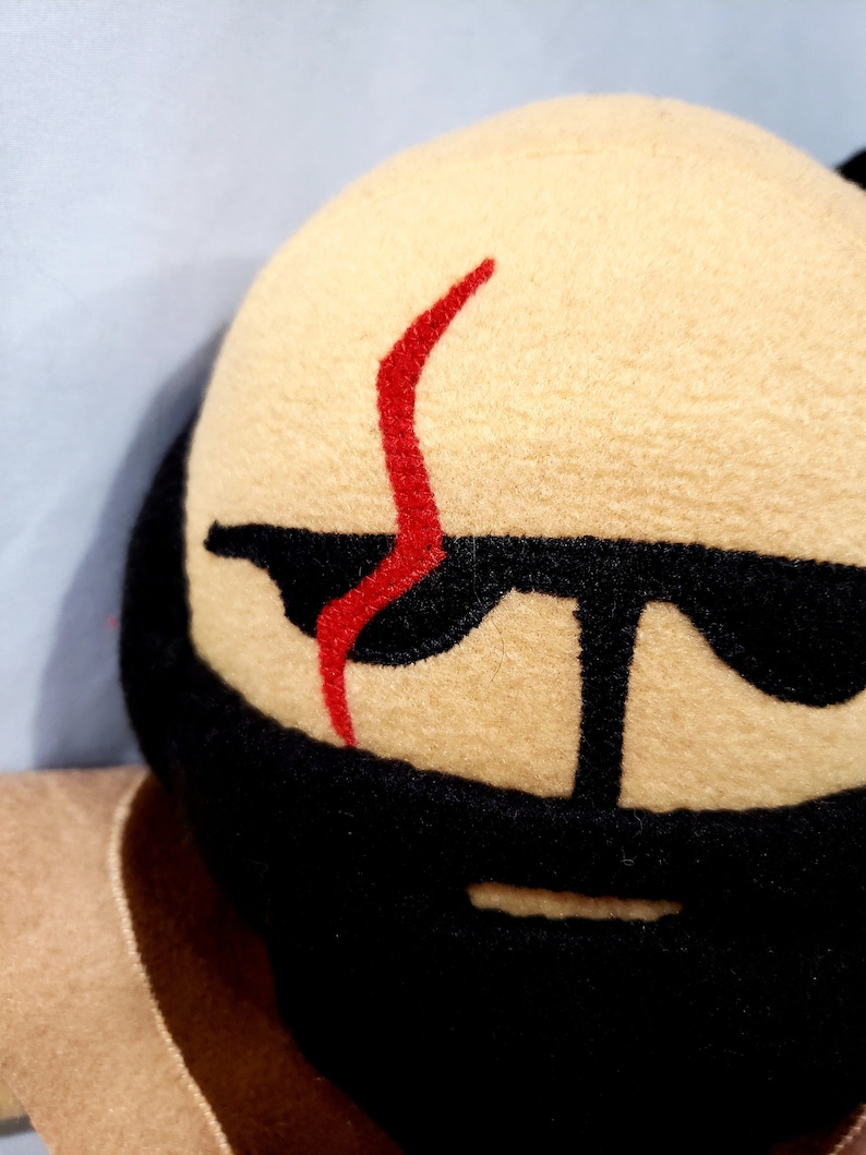 May include: A plush toy with a tan face, black beard, and red scar. The toy has black eyes and a black mouth.