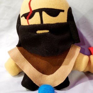 May include: A plush toy with a tan face, black beard, and brown and tan clothing. The toy has a red scar on its face and is wearing sunglasses. A blue ball is in front of the toy.