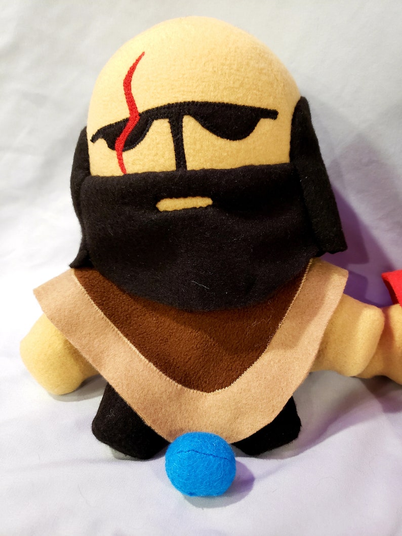 May include: A plush toy with a tan face, black beard, and brown and tan clothing. The toy has a red scar on its face and is wearing sunglasses. A blue ball is in front of the toy.