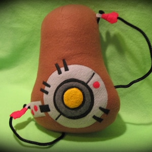 Puede incluir: Un juguete de peluche marrón con forma de pera con un círculo blanco y un centro amarillo en la parte delantera. El juguete tiene un cordón negro con un lazo rojo unido a la parte superior.