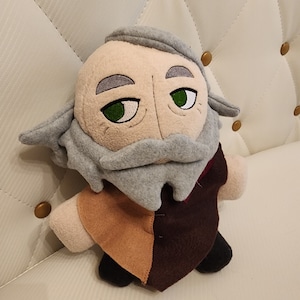 May include: A plush toy of an older person with a gray beard and brown and tan clothing. The toy has green eyes and a white face.