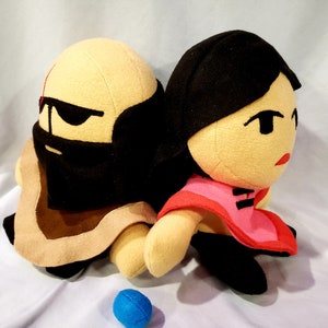 May include: Two plush toys, one with a brown robe and black eye mask, the other with a red and black dress, sitting side by side.