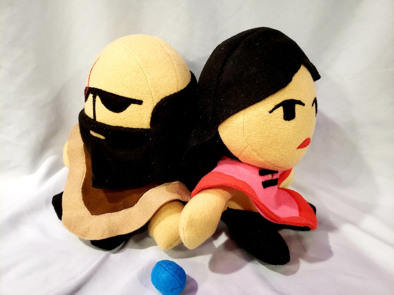 May include: Two plush toys, one with a brown robe and black eye mask, the other with a red and black dress, sitting side by side.