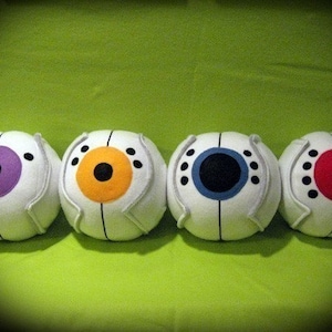 May include: Four plush white eye-shaped toys with different colored irises. The irises are purple, orange, black, and red.