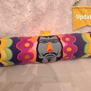 May include: A cylindrical decorative pillow with a cartoon face design. The pillow is primarily purple with colorful accents of orange, pink, yellow, green, and blue. A small yellow banner with the word "Updated!" hangs in the background.