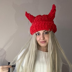 Pode incluir: Um chap&eacute;u vermelho de croch&ecirc; com chifres de diabo e uma corrente prateada. O chap&eacute;u &eacute; usado por uma pessoa com cabelo longo e loiro e um su&eacute;ter branco.