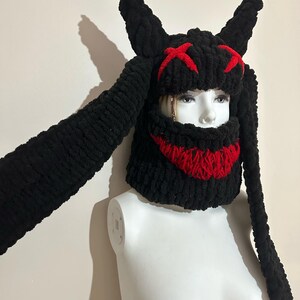 May include: Black knitted hat with long bunny ears and a red stitched smile and eyes. The hat is on a white mannequin.