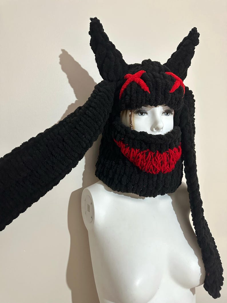 May include: Black knitted hat with long bunny ears and a red stitched smile and eyes. The hat is on a white mannequin.