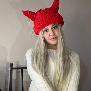 Pode incluir: Um chap&eacute;u vermelho de croch&ecirc; com chifres e uma corrente prateada. O chap&eacute;u est&aacute; sendo usado por uma pessoa com cabelo loiro comprido.