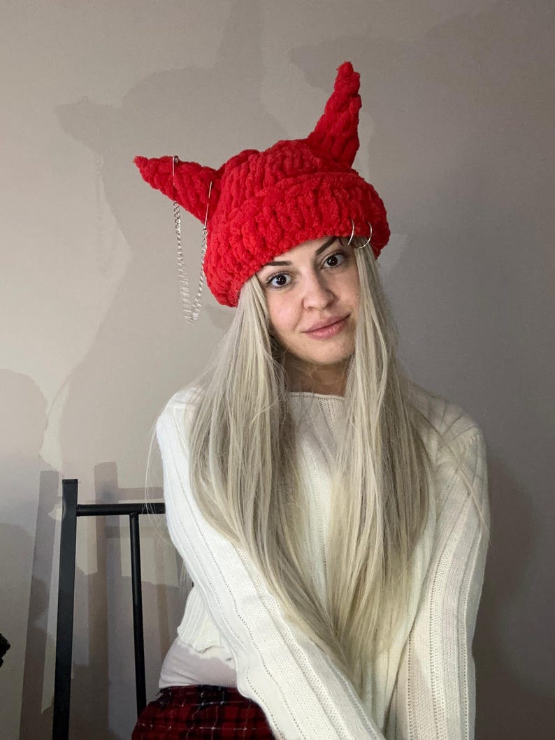 Pode incluir: Um chap&eacute;u vermelho de croch&ecirc; com chifres e uma corrente prateada. O chap&eacute;u est&aacute; sendo usado por uma pessoa com cabelo loiro comprido.